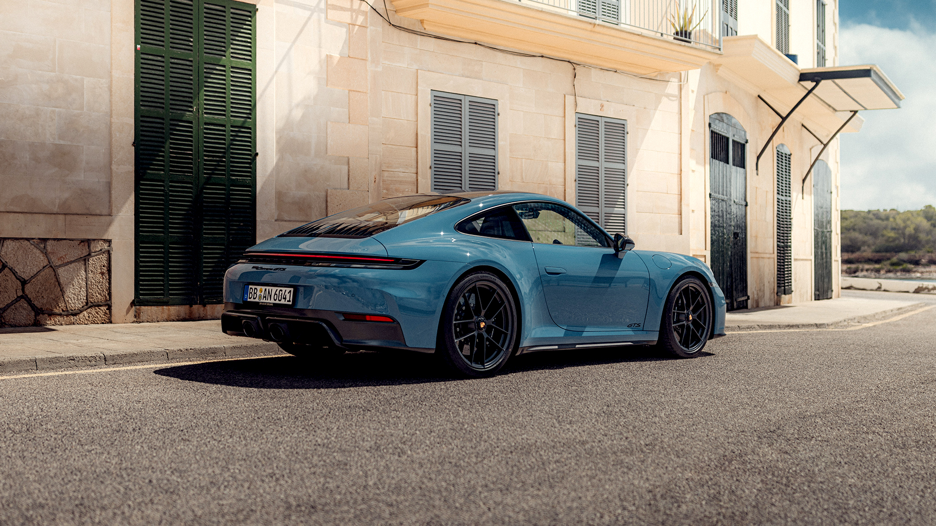 Rear three-quarter view of a light blue Porsche 911 Carrera GTS parked on a Mediterranean-style cobblestone street, featuring black center-lock wheels, quad exhaust outlets, illuminated taillight strip, and GTS badging